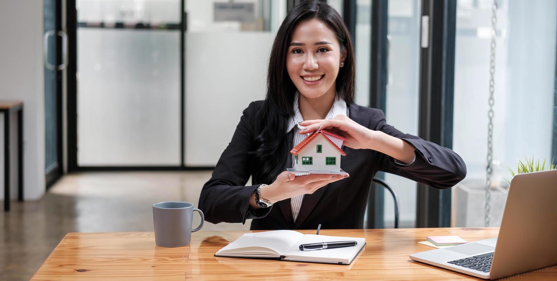 miniatuur huis in de handen van een aziatische vrouw makelaar woningkrediet werken op kantoor. naar de camera kijken. foto