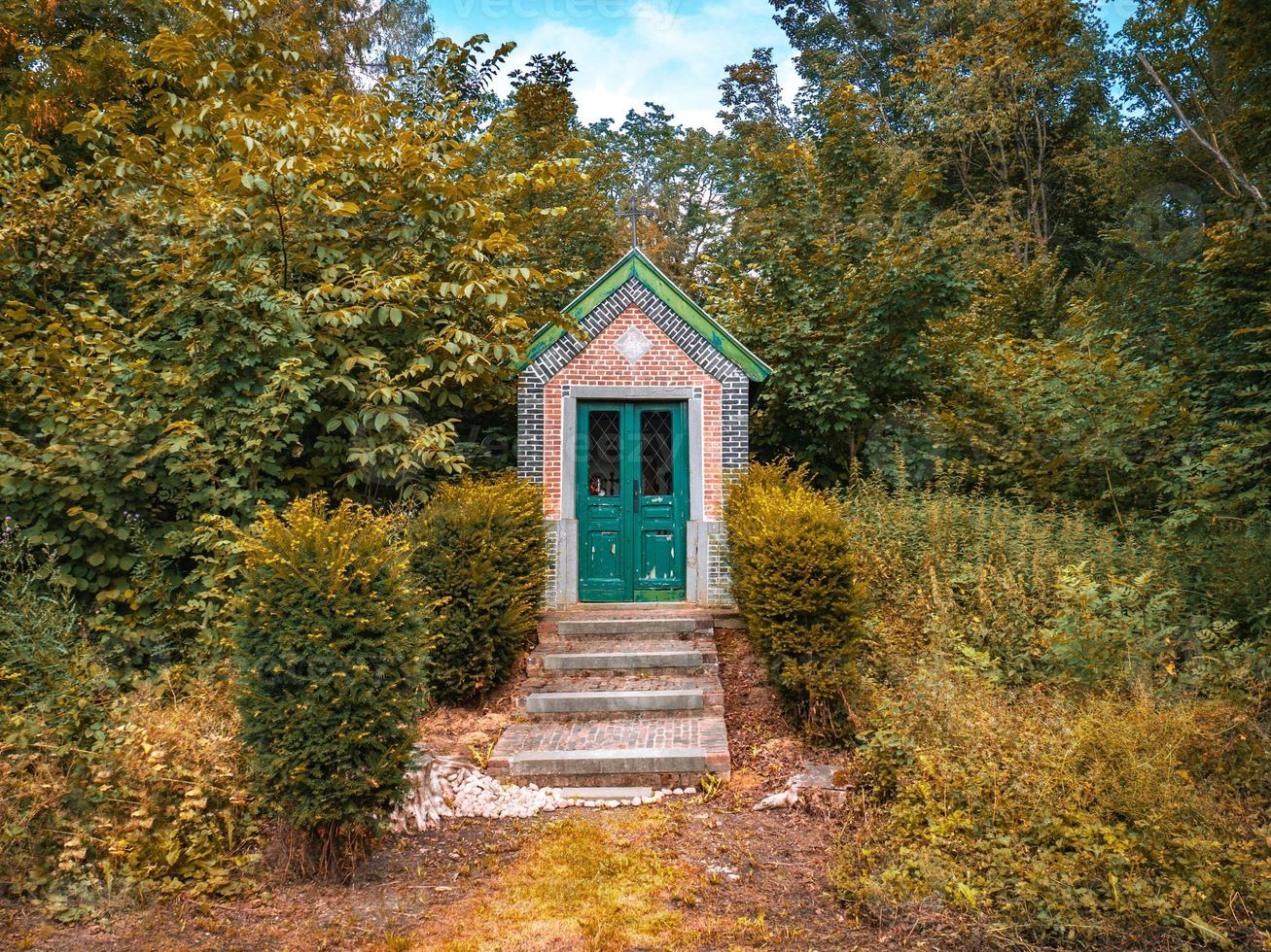 een historische kleine kapel met zijn groene deur en dak, rode en blauwe bakstenen en stenen trappen te midden van groen gras en bomen in Tervuren, België. foto