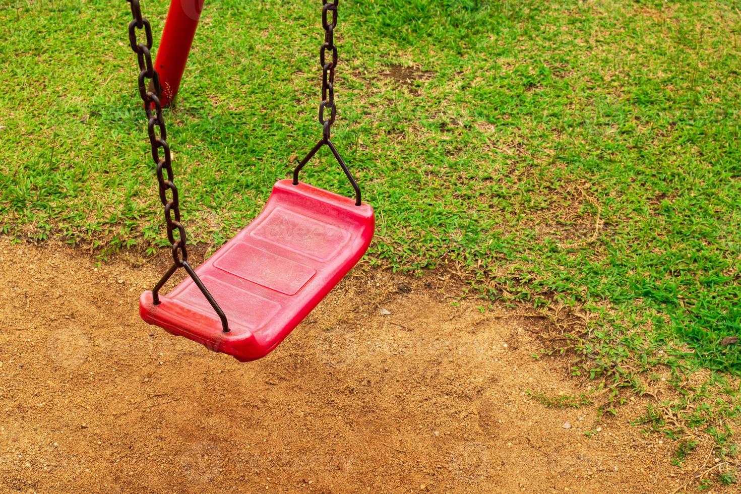 een rode schommel is bevestigd aan een roestige oude ketting in de speeltuin. in het park zag de grond en het gras foto