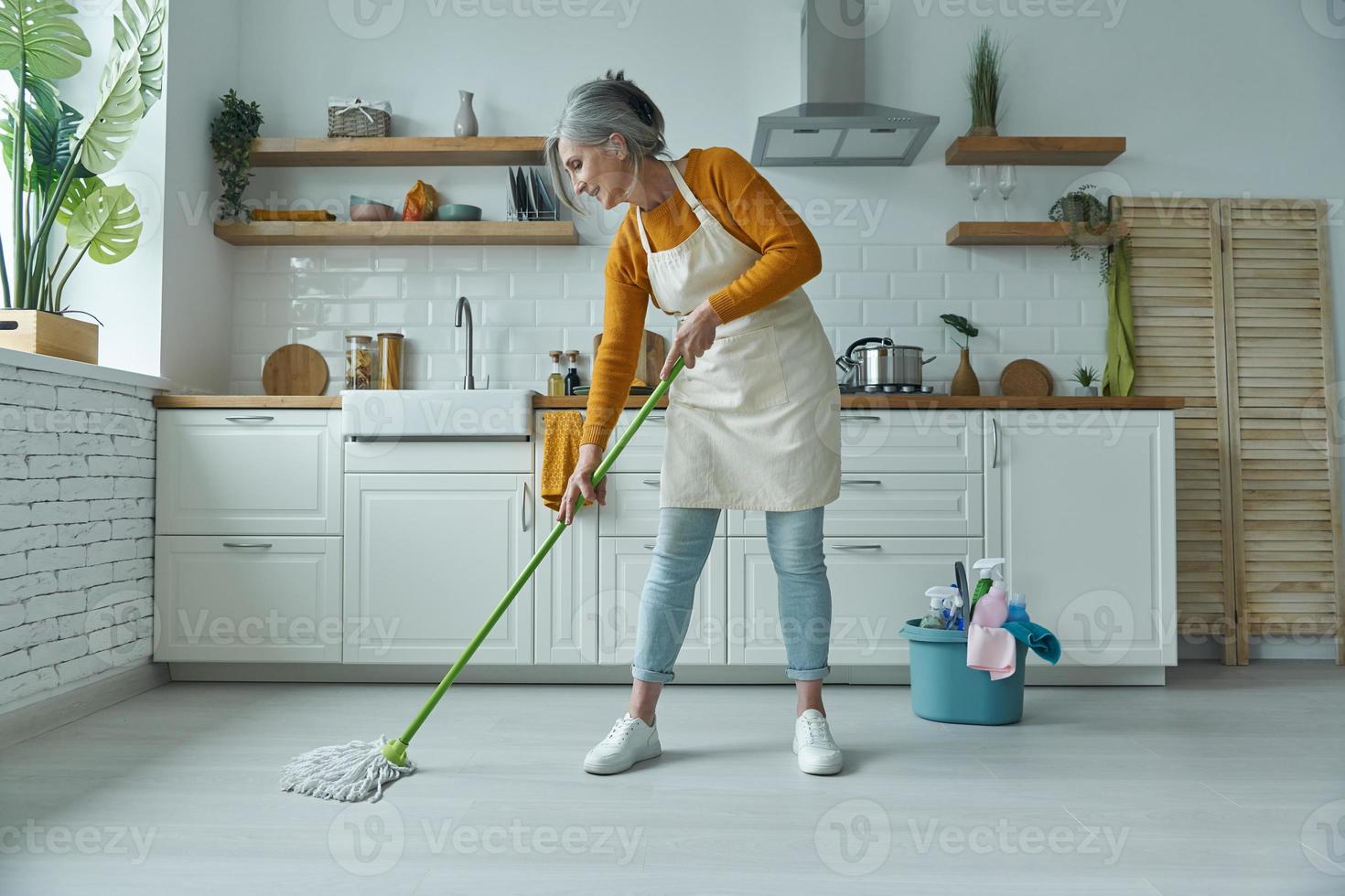 volledige lengte van zelfverzekerde senior vrouw die de vloer schoonmaakt met dweil terwijl ze in de huishoudelijke keuken staat foto