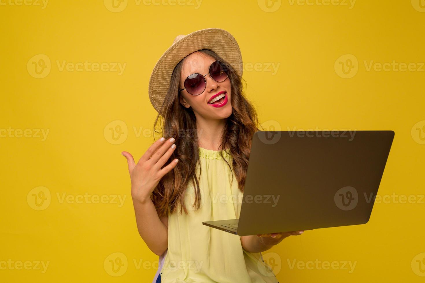 stijlvol trendy meisje met golvend haar in zonnebril en hoed doet een videogesprek met laptop met vrolijke emoties foto