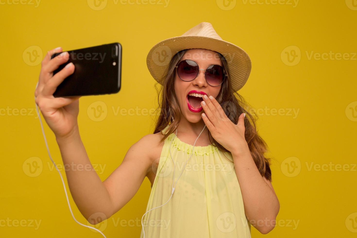 opgewonden mooi meisje met lang golvend haar in zonnebril en hoed bedekt lippen met hand en maakt selfie op smartphone foto