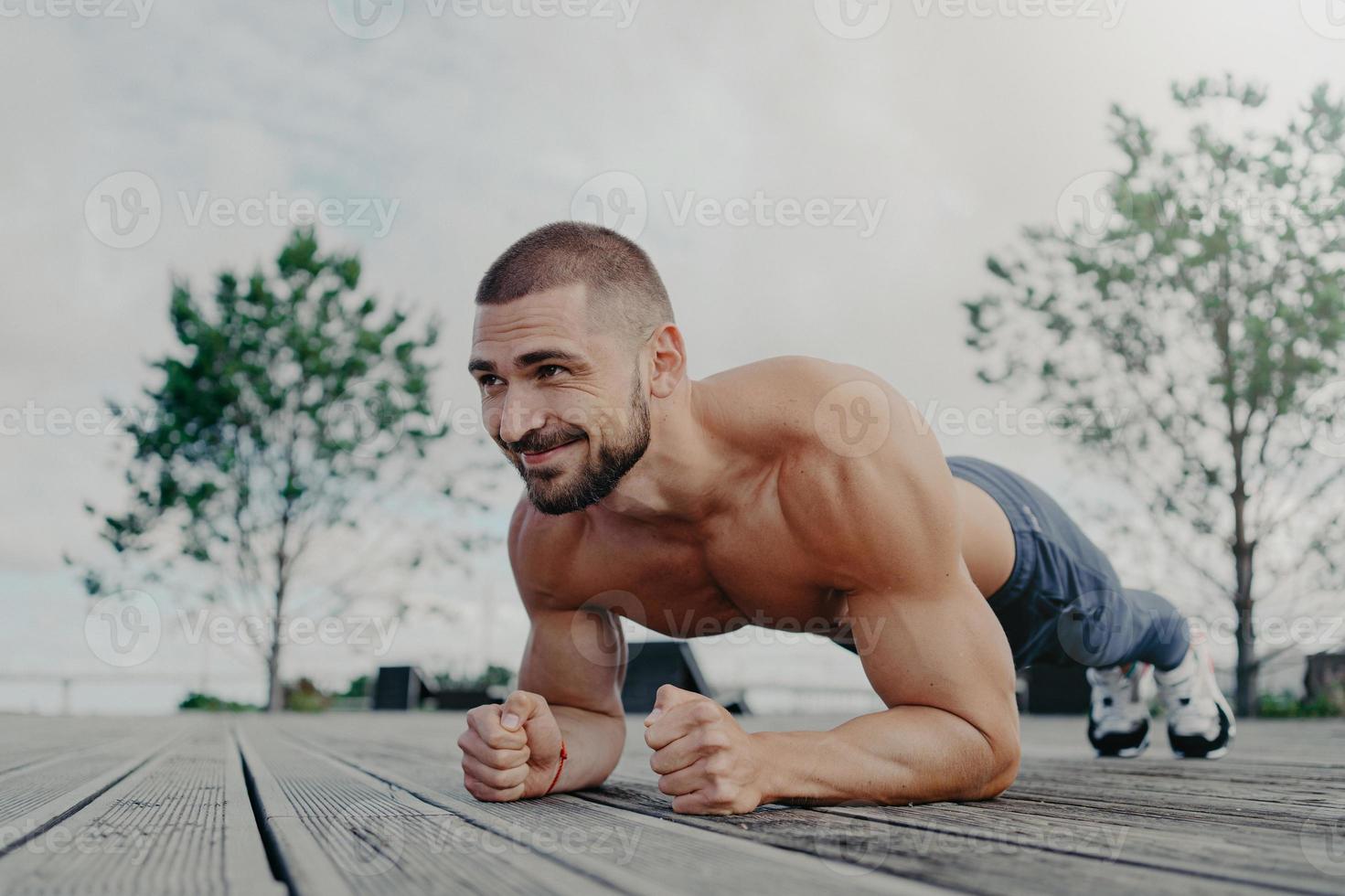 shirtless vastberaden bebaarde man met naakte torso staat in plank pose en glimlacht blij, demonstreert uithoudingsvermogen, poseert buiten, denkt aan zijn lichaam en gezondheid. atletische man traint buiten foto