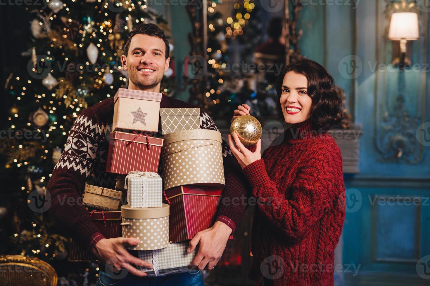 lachende vrouw draagt rode gebreide trui houdt glazen bol vast als sierboom, staat in de buurt van haar man die stapels cadeautjes vasthoudt, gaat ze presenteren aan vrienden en familieleden. druk stel foto