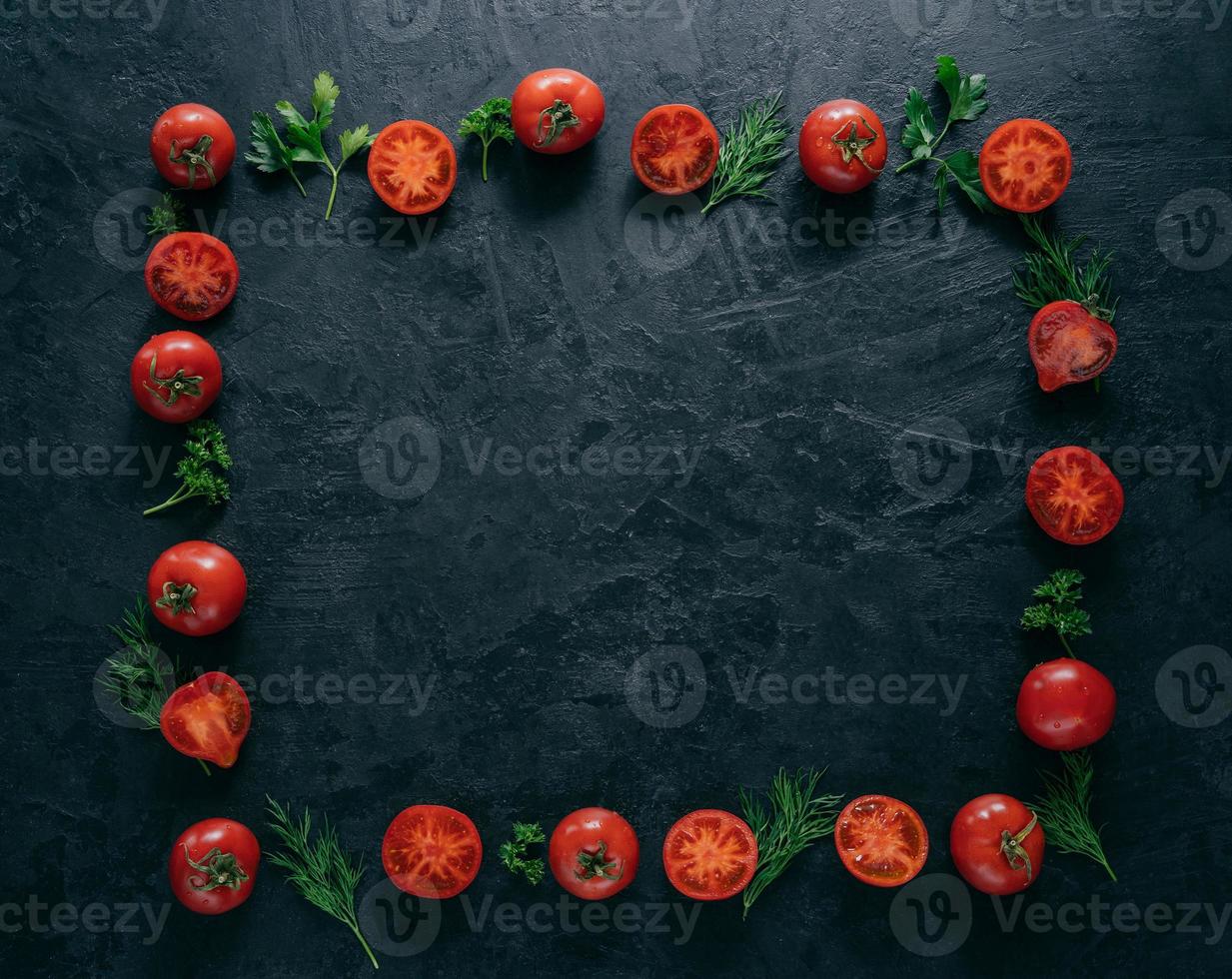 plat leggen van rode rijpe tomaten ligt in de vorm van een frame op een donkere achtergrond met groene peterselie en dille. gezond vegetarisch gerecht. horizontaal schot. kopieer ruimte voor tekst foto