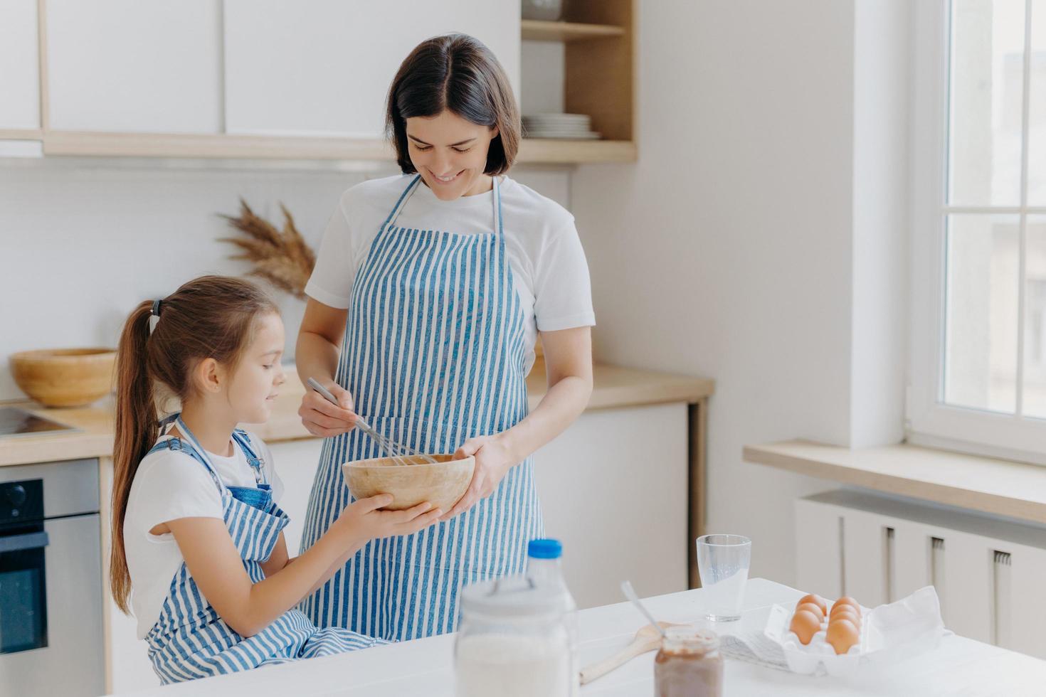 gelukkige familie bij keuken. lieve vrouw en haar dochter bereiden samen de bakkerij, dragen schorten, houden van samen koken, genieten van huiselijke sfeer, hebben plezier binnen. kinderen, moederschap, bakconcept foto