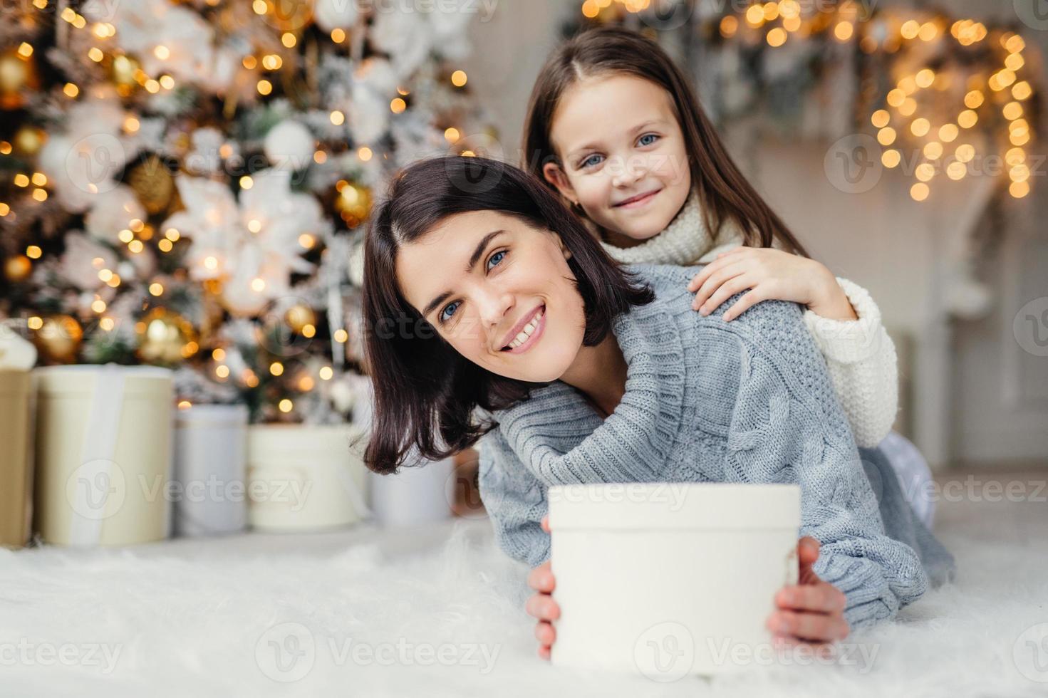 kinderen, familie en viering concept. schattige vrouw in gebreide trui houdt witte geschenkdoos vast en klein kind staat achter haar rug, heeft een goed humeur voor oudejaarsavond. wachtend wonder foto