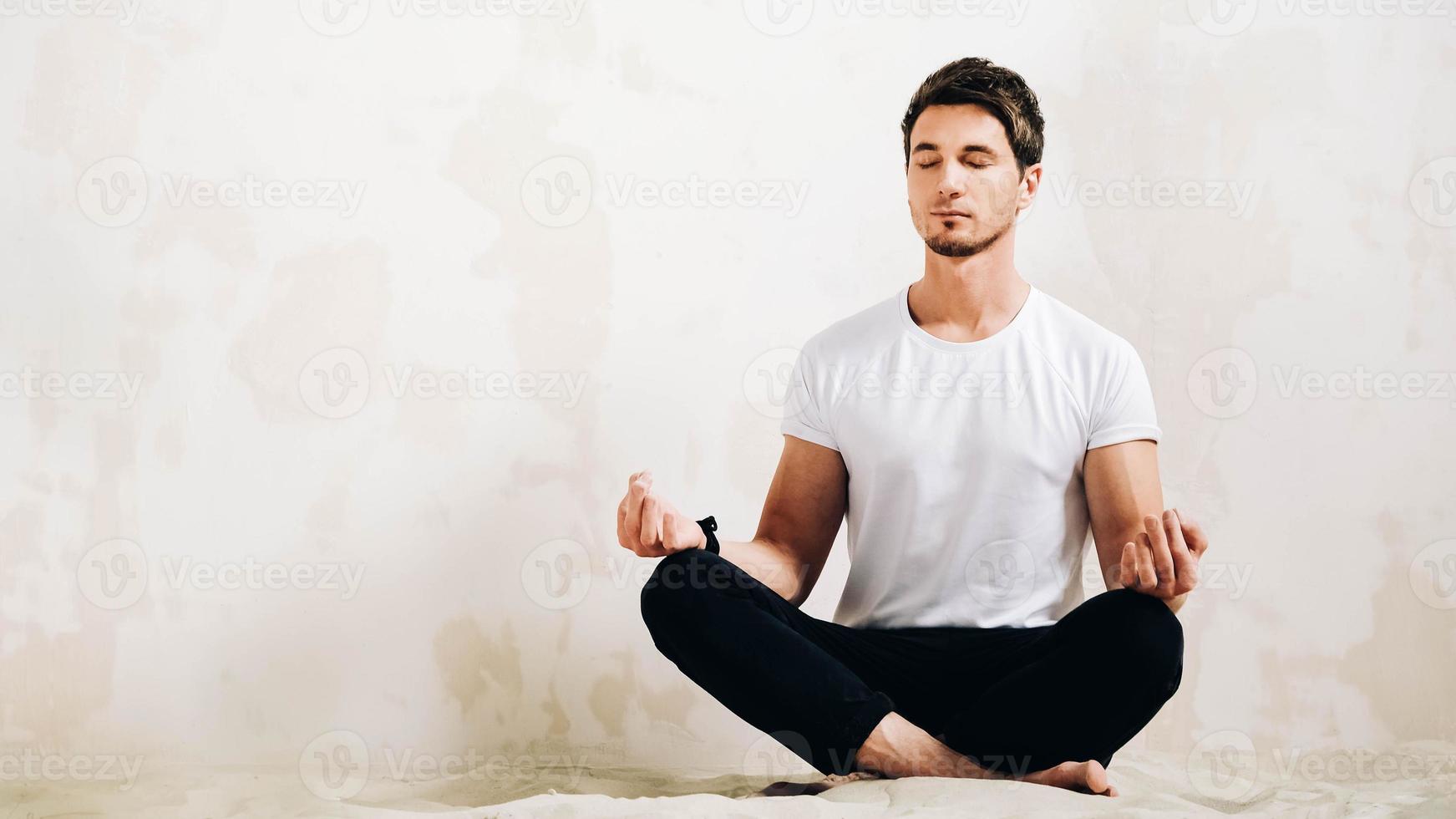 jonge man zit in een meditatieve pose op zand tegen de achtergrond van een muur foto