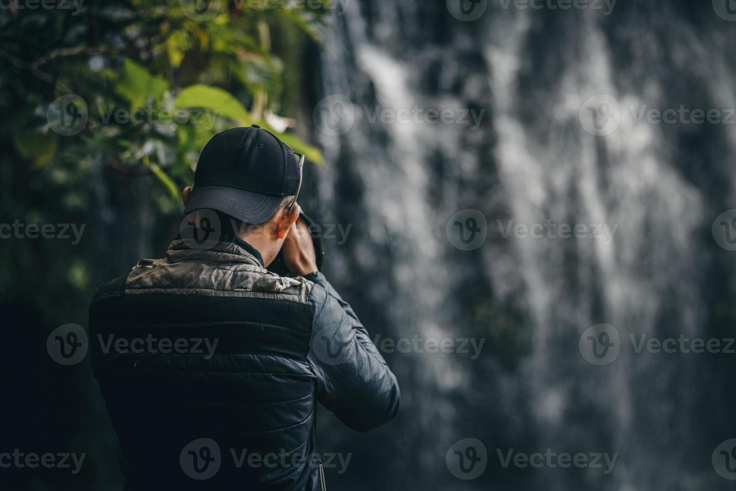 close-up van de achterkant fotograaf in casual stijl draag jas en hoed maakt gebruik van moderne camerafotografie met wazige waterval. foto