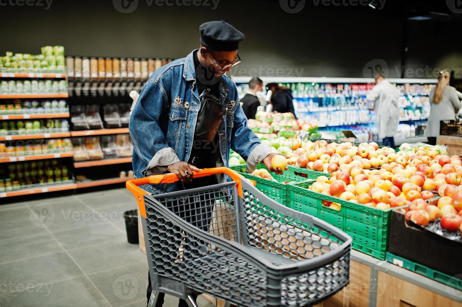 stijlvolle casual Afro-Amerikaanse man bij jeans jasje en zwarte baret bij biologische sectie van supermarkt met winkelwagentje kar. foto