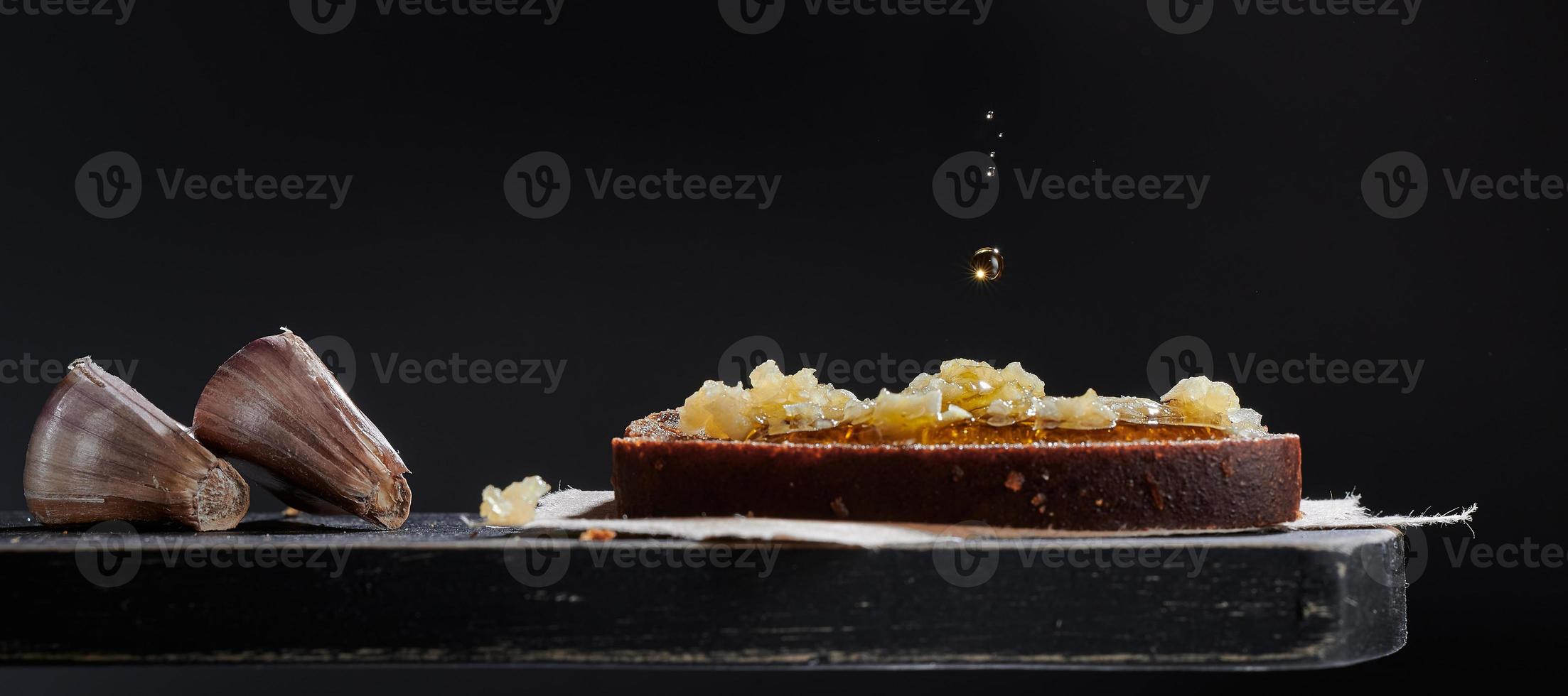 een stuk zwart brood met knoflook op een houten bord. een druppel olijfolie valt op de boterham. donkere rustieke achtergrond. foto