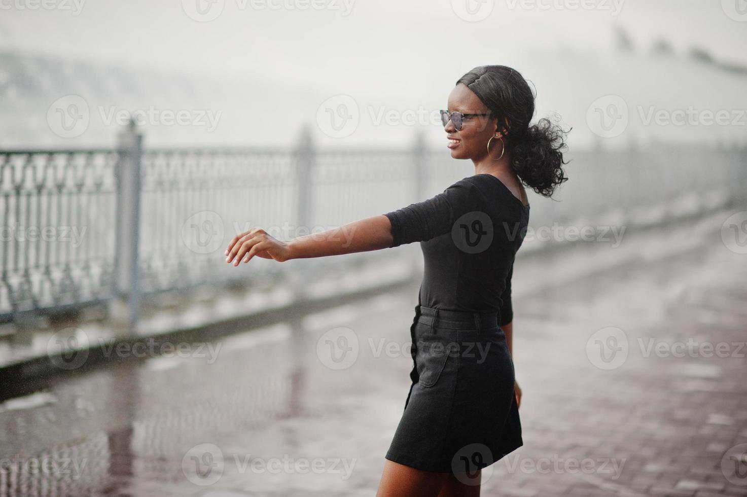 afro-amerikaans meisje in zonnebril, zwarte kleding en shirt poseerde buiten. modieuze zwarte vrouw tegen regen van fonteinen. foto