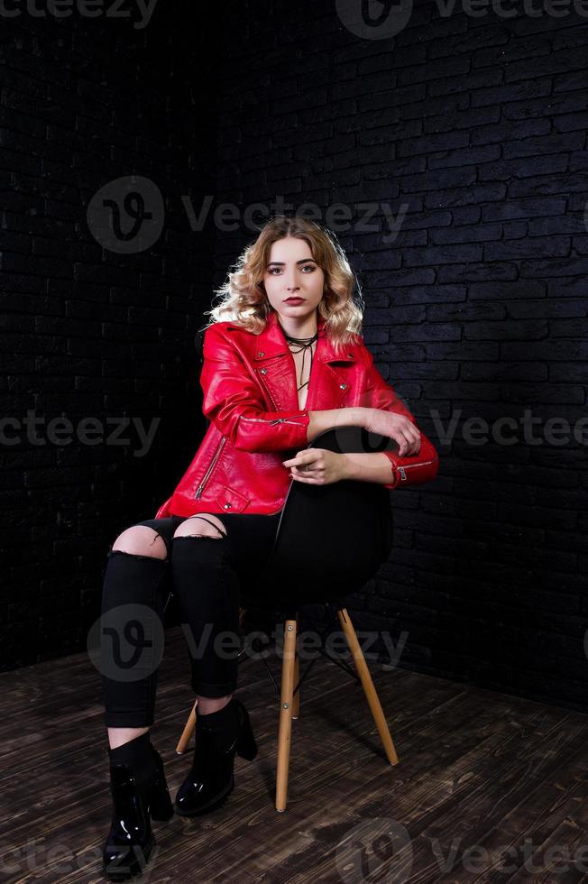 studio portret van blond meisje in rode leren jas gesteld op stoel tegen bakstenen muur. foto