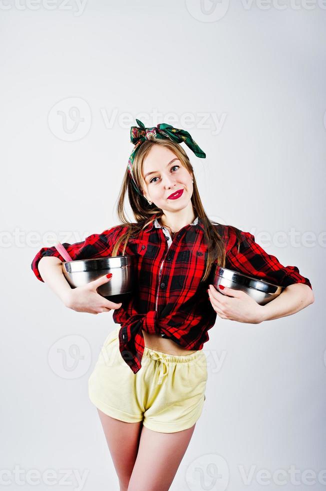 jonge grappige huisvrouw in geruit overhemd en gele korte broek pin-up stijl met twee steelpan geïsoleerd op een witte achtergrond. foto