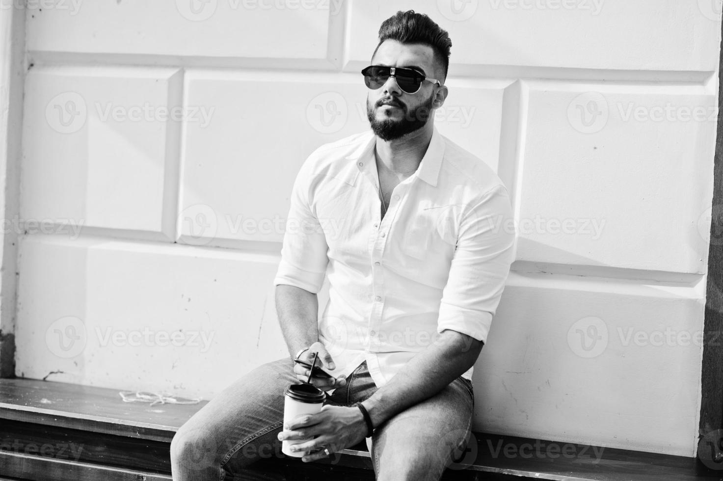 stijlvolle lange Arabische man model in wit overhemd, jeans en zonnebril poseerde op straat van de stad en houd mobiele telefoon bij de hand. baard Arabische man met kopje koffie zitten en rusten. foto