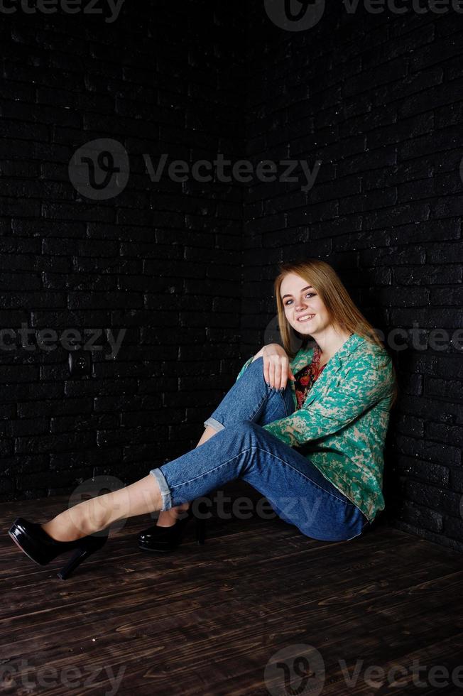 stijlvol blond meisje in jas en spijkerbroek tegen zwarte bakstenen muur in de studio. foto