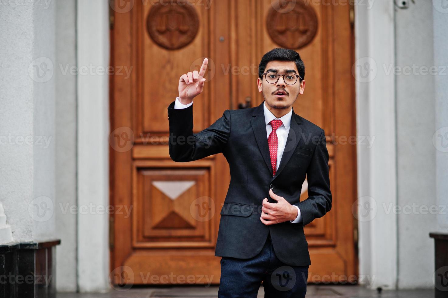 indiase jonge man met een bril, draag een zwart pak met rode stropdas die buiten tegen de deur van het gebouw staat, laat de vinger zien, hij heeft een idee. foto