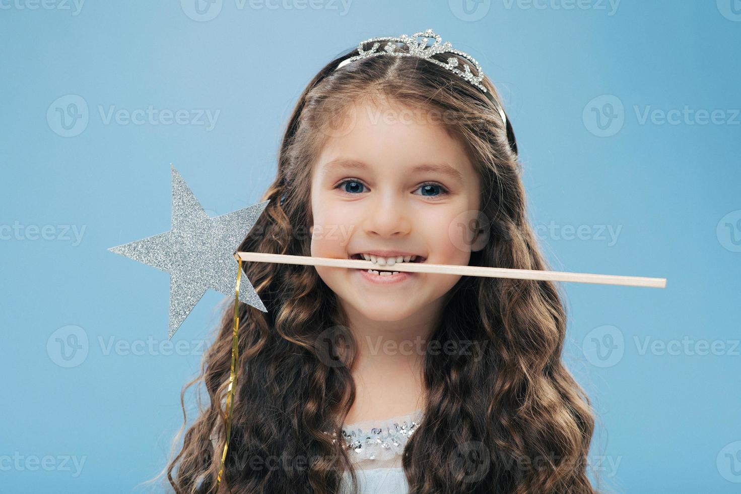 horizontale opname van een aantrekkelijk lachend klein kind houdt een toverstaf in de mond, heeft bruin krullend haar, poseert over een blauwe achtergrond, kijkt rechtstreeks naar de camera, draagt een kroon. kinderen en magisch concept foto