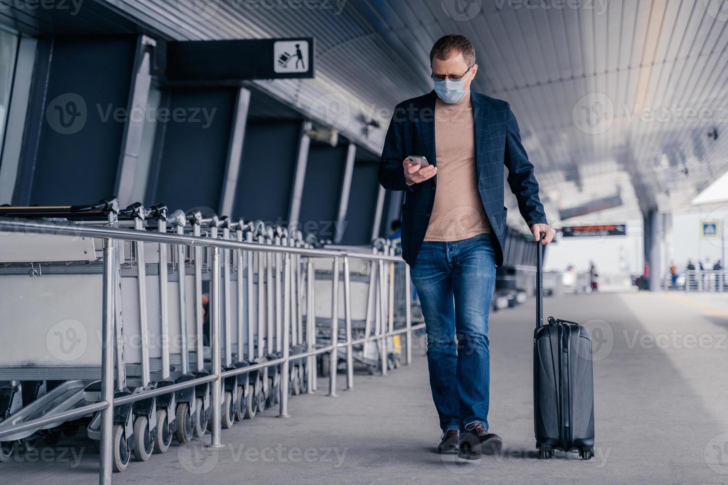 mannelijke passagier loopt op luchthaven, houdt koffer vast, controleert nieuwsfeed op smartphone, draagt wegwerp medisch masker tijdens pandemisch coronavirus, probeert veilig te zijn tijdens ziektevirus. ademhalingssymptomen foto