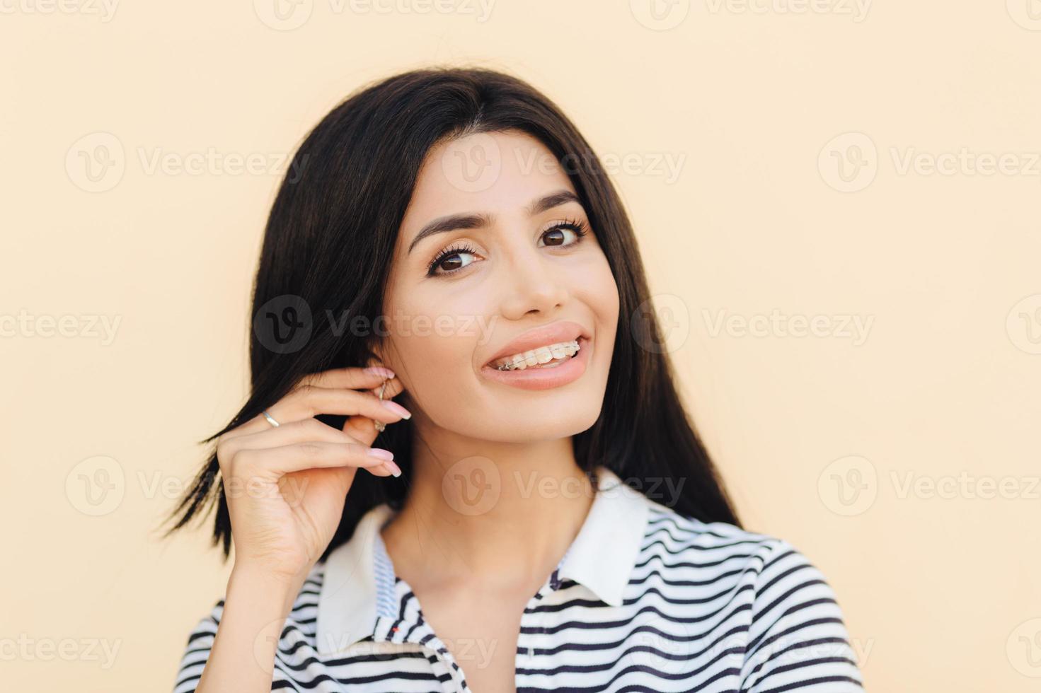 headshot van schattige mooie vrouw heeft een brede glimlach, donker haar, houdt de hand op het oor, toont witte tanden met beugels, kijkt direct in de camera, heeft een gelukkige uitdrukking, geïsoleerd over studio-achtergrond. foto