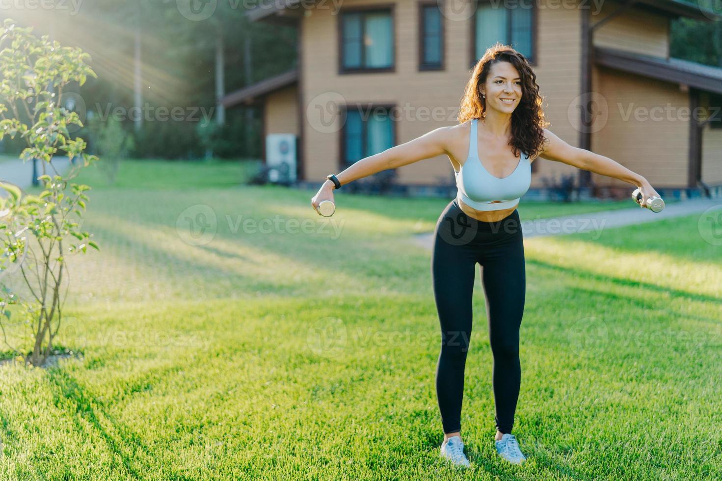 slanke gemotiveerde brunette vrouw gekleed in bijgesneden top en legging, heeft training met halters, poseert op groen gazon in de buurt van privéhuis, heeft een perfecte lichaamsvorm. gezonde levensstijl en sportconcept foto