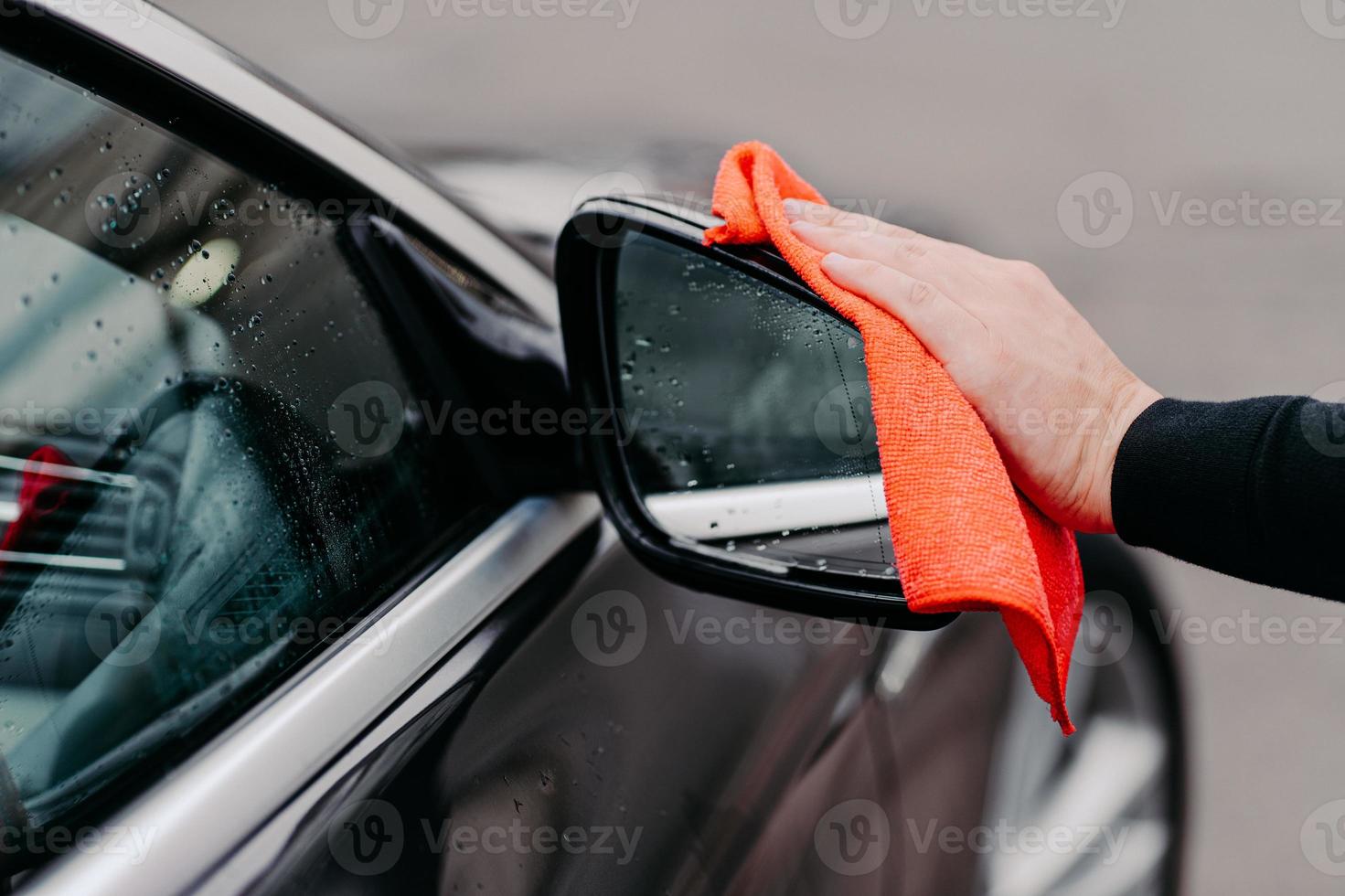 close-up van mans hand die water afveegt op zwarte auto met microvezeldoek. focus op auto zijspiegel. zelfbediening voor vervoer foto