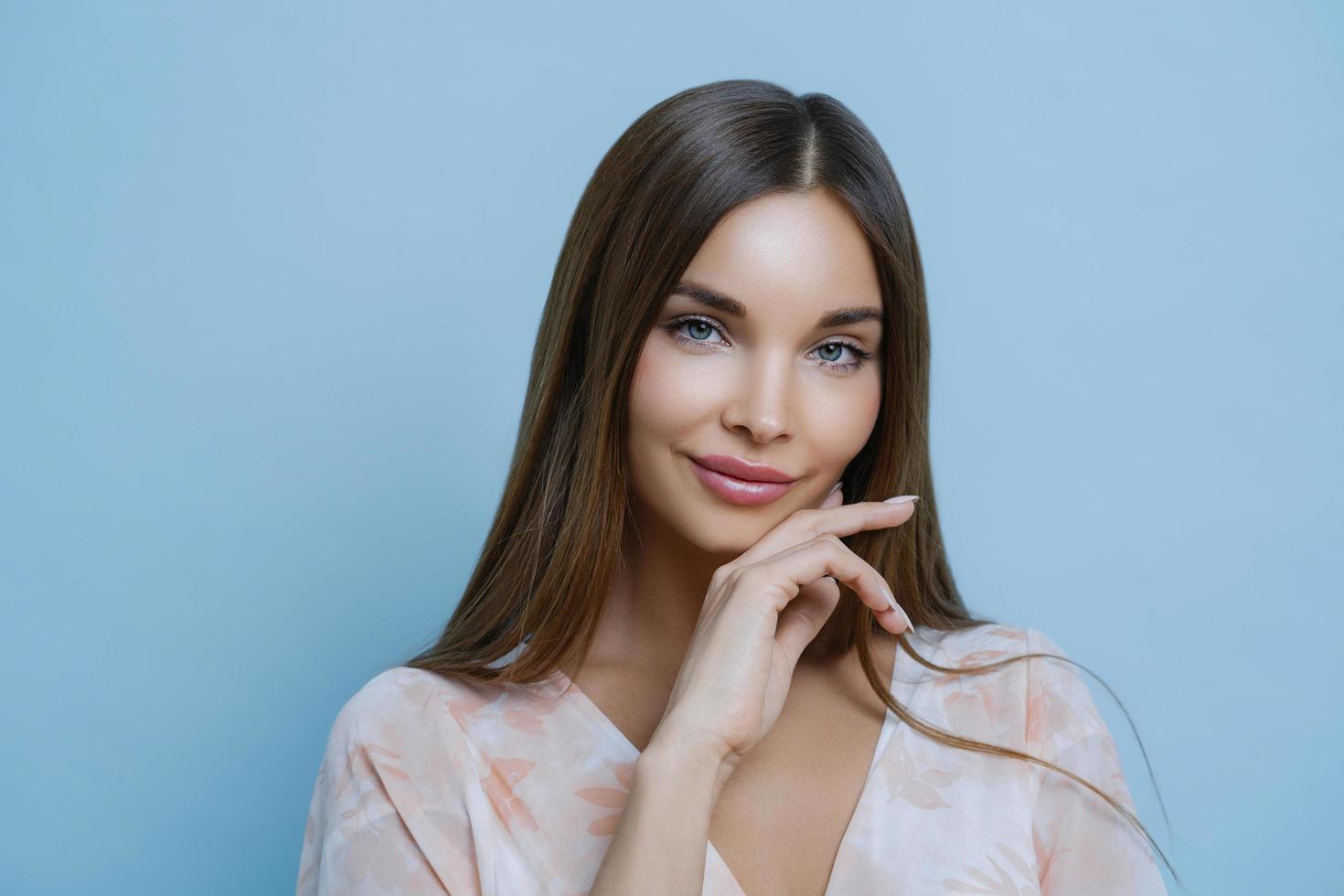 verleidelijke aangenaam ogende vrouw met lang donker steil haar raakt kaaklijn aan, kijkt uit naar speciale gebeurtenis, neemt deel aan professionele fotoshoot, heeft een gezonde schone huid, poseert over blauwe achtergrond foto