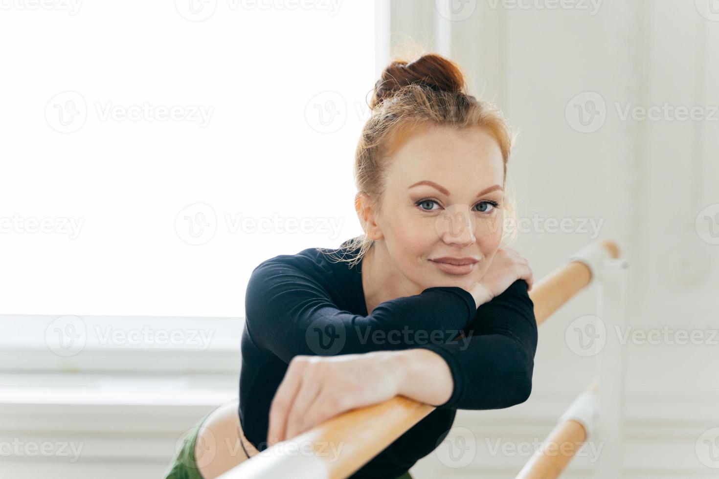 headshot van rustige mooie vrouwelijke balletdanser met gemberbroodje, draagt sportkleding, heeft een flexibel lichaam, poseert in de buurt van bars, ontwikkelt dansvaardigheden, heeft minimale make-up. professionele ballerina in studio foto