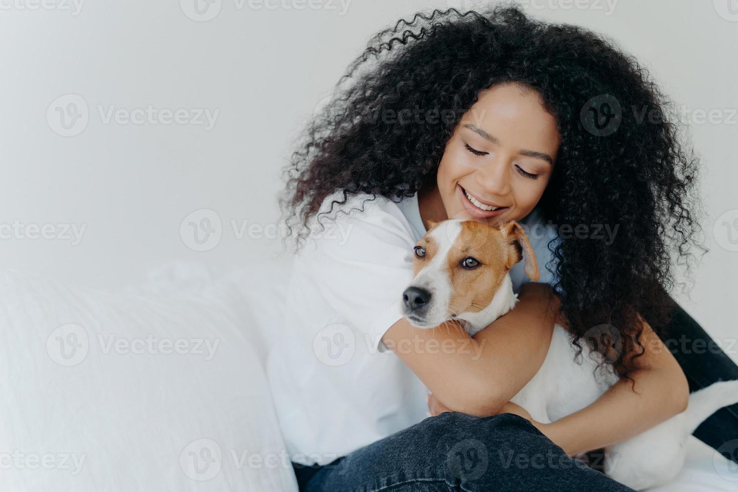 gelukkige afro-amerikaanse vrouw drukt liefde uit voor de hond, omarmt huisdier, gekleed in vrijetijdskleding, zit op een comfortabel bed in de slaapkamer, slaapt met huisdier. jack russell terrier met eigenaar in de ochtend foto