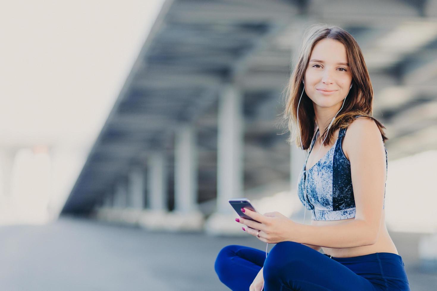 tevreden vrouw met een gezonde huid, voelt zich verfrist terwijl ze naar favoriete nummer luistert in oortelefoons, gekleed in sportkleding, zit in lotushouding, wacht op vriend, poseert buiten. mensen en levensstijl concept foto