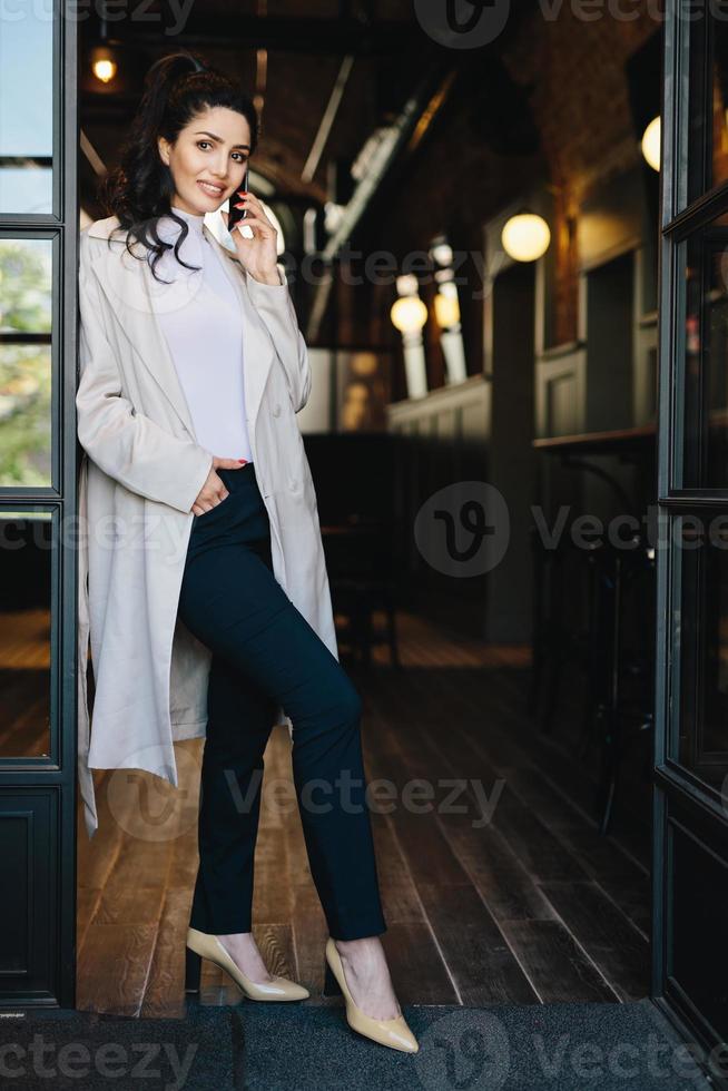 stijlvolle jonge vrouw met donker haar, gekleed in een witte jas, zwarte broek en schoenen met hakken poseren in de camera terwijl ze tegen de achtergrond van een café staat te chatten via de telefoon met de hand in de zak foto