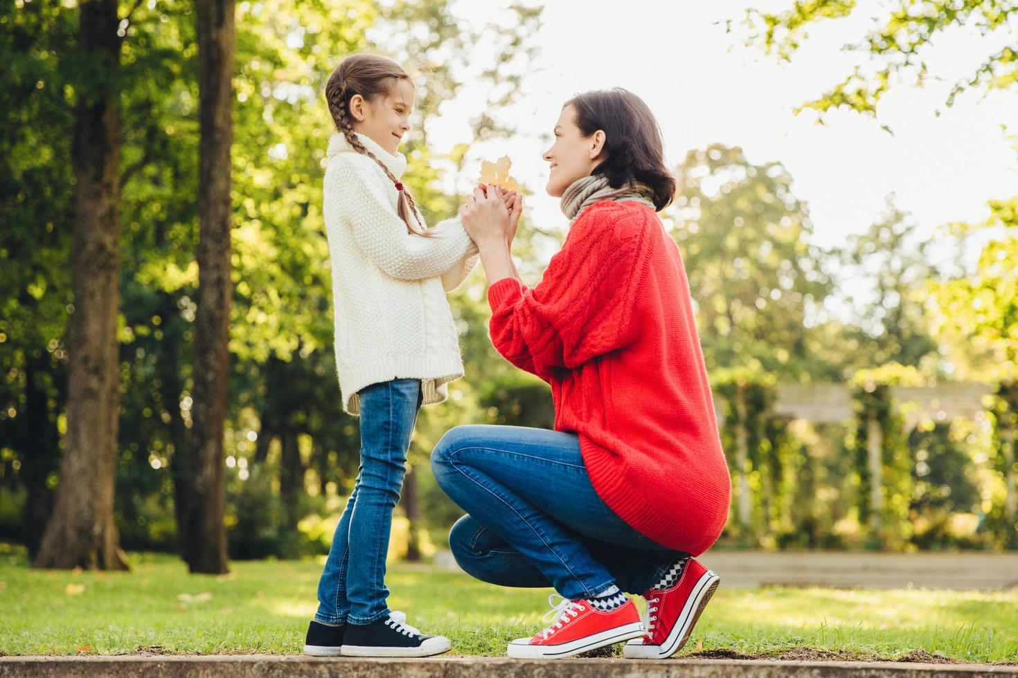 jonge moeder in warme gebreide rode trui speelt met haar kleine dochter in het park, geeft haar blad, geniet van zonnig herfstweer. aanhankelijke moeder en klein kind brengen tijd samen buiten door foto