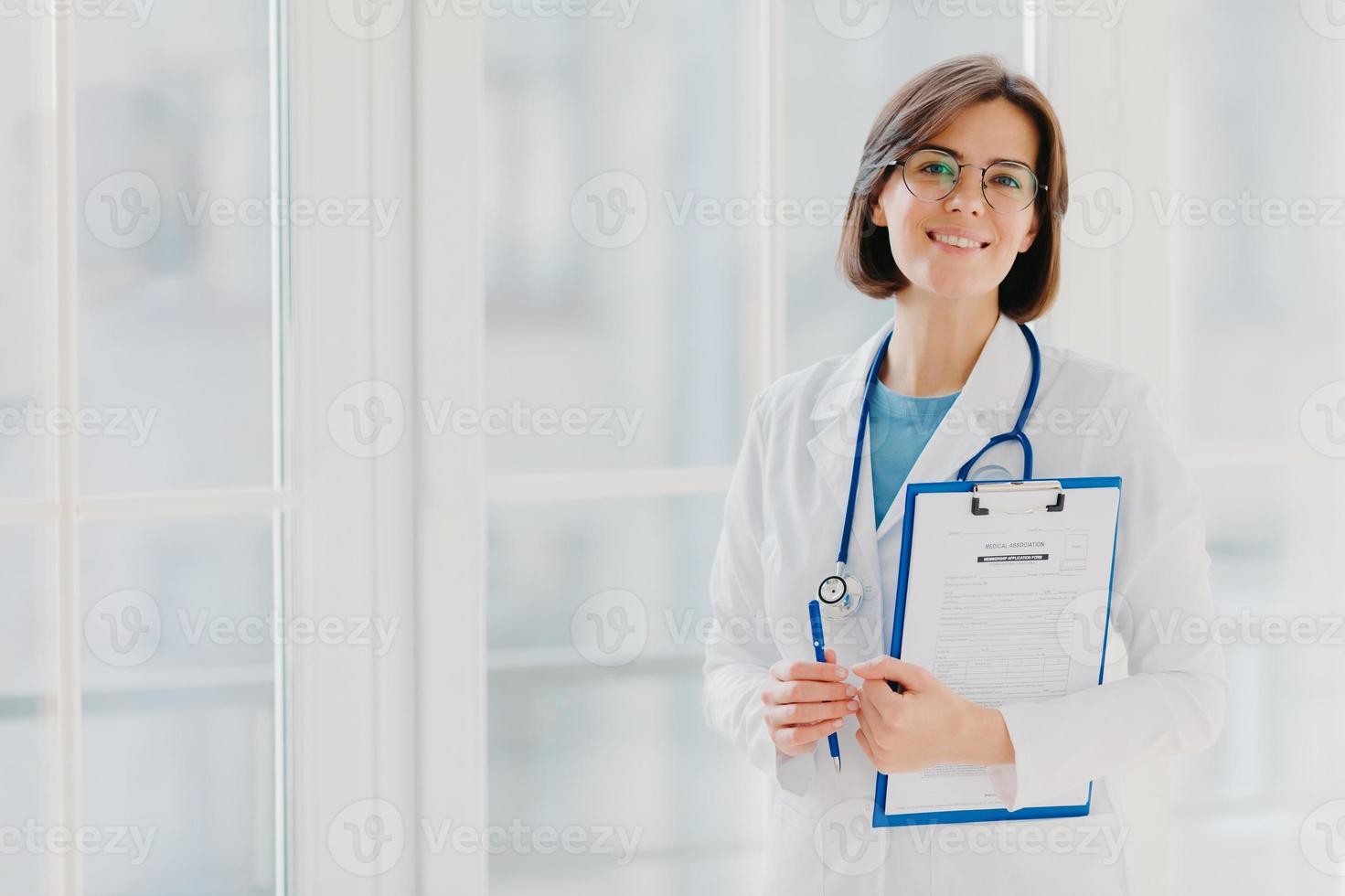 horizontaal schot van vrouw arts staat met klembord, vult aanvraagformulier, houdt pen vast, glimlacht positief, geniet van haar werk, helpt mensen, staat in witte medische jurk tegen groot raam foto