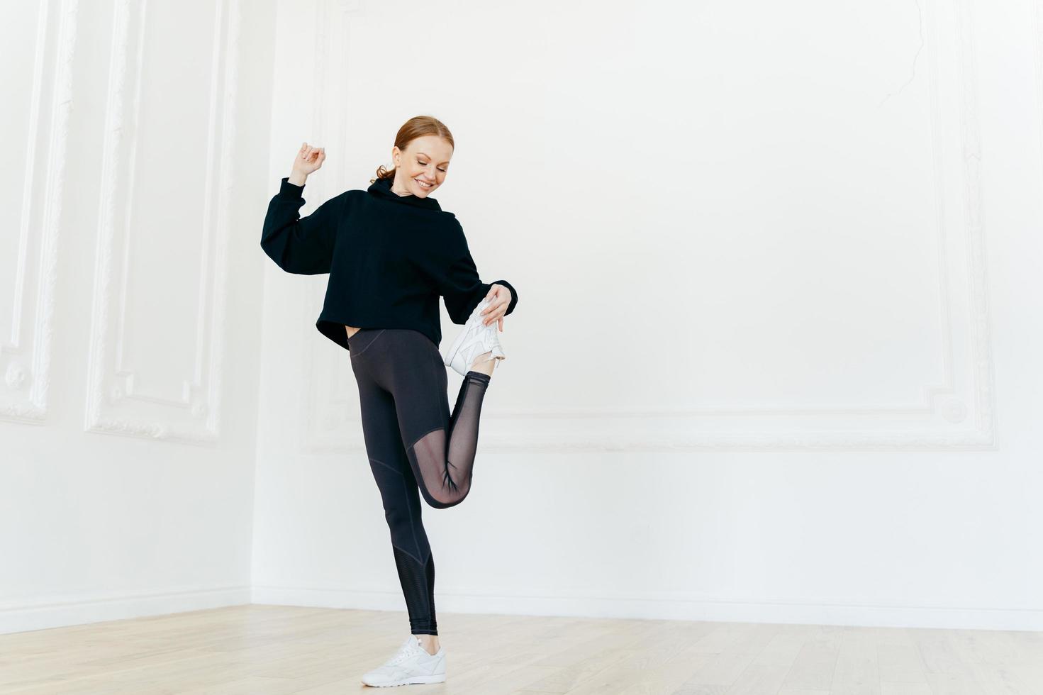 volledige lengte shot van tevreden slanke jonge vrouw heeft make-up staat op één been, heft voeten op, gekleed in zwarte sportkleding, heeft een gezonde lichaamsvorm, blije uitdrukking, toont mooie soepelheid. foto