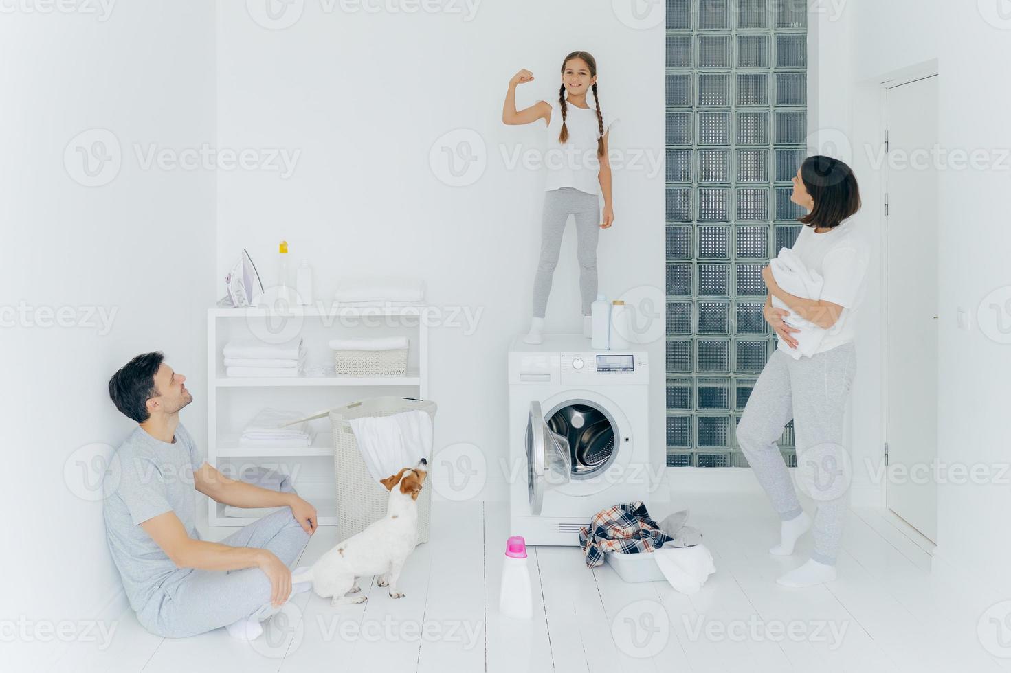 sterk vrolijk meisje heft arm op, toont biceps, amuseert ouders, pauze na het wassen van kleding, poseert in ruime witte wasruimte met huisdier, wasmachine en schoonmaakmiddelen. gezin en huishoudelijk werk foto