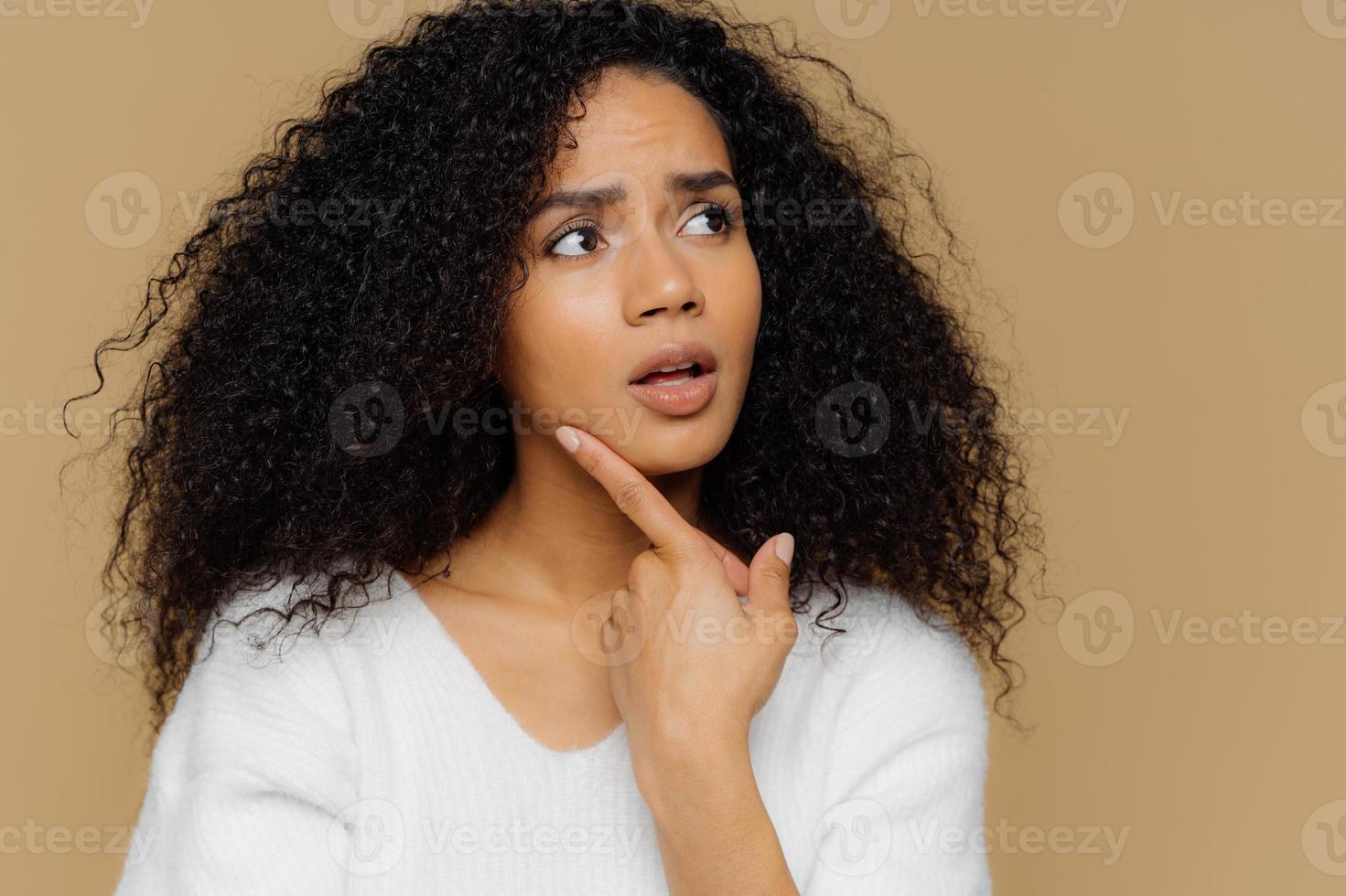 verbaasd vrouwelijk model houdt de vinger op de kin, kijkt bedachtzaam opzij, heeft gefrustreerde gezichtsuitdrukking, krullend afro-haar, draagt een casual witte trui, geïsoleerd over bruine achtergrond. Close-up shot foto