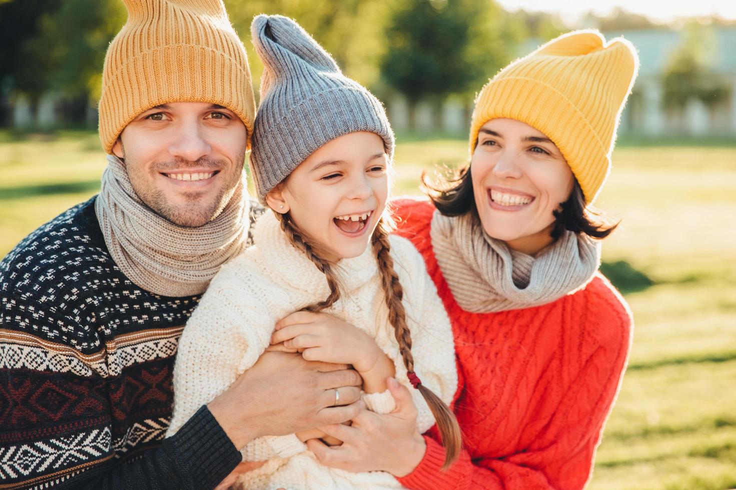 speels klein kind met staartjes draagt warme kleding, brengt vrije tijd door met lieve aanhankelijke ouders, heeft een gelukkige uitdrukking, voelt zich ontspannen. drie jonge gezinsleden genieten van aangename momenten foto