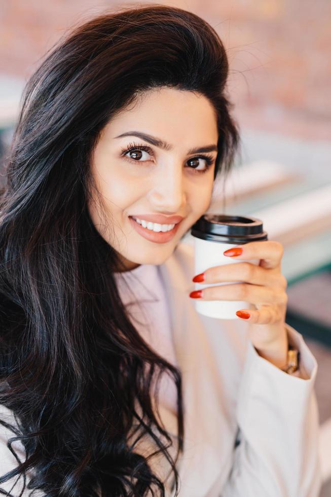 schoonheid, jeugd, lifestyle en ontspanning concept. jonge brunette vrouw met goed gevormde dunne wenkbrauwen, gezonde huid en rode manicure met afhaalkoffie en glimlachend in de camera kijkend foto