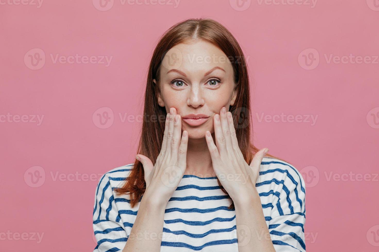headshot van sproeten mooie vrouw houdt handen in de buurt van lippen, heeft minimale make-up, gezonde huid, bruin haar, toont natuurlijke schoonheid, gekleed in gestreepte outfit, geïsoleerd op roze achtergrond. foto