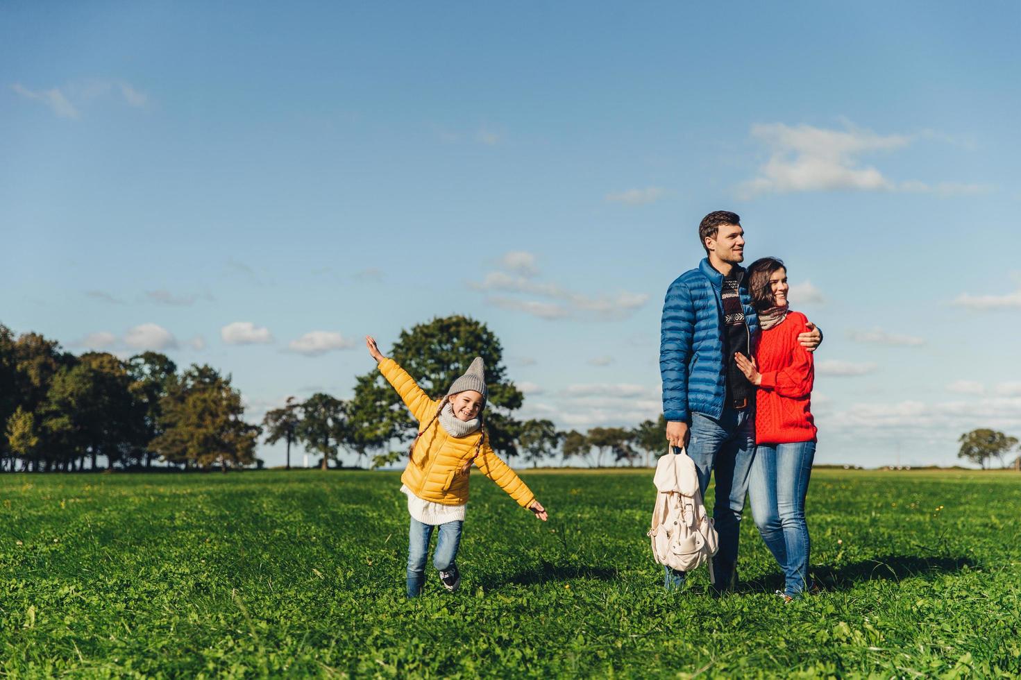 speels klein kind rent op groene weide, speelt in de buurt van ouders, die elkaar omhelzen, in de verte kijken met doordachte uitdrukkingen. familie heeft wandelen of wandelen op het platteland, rust na lawaai stad foto