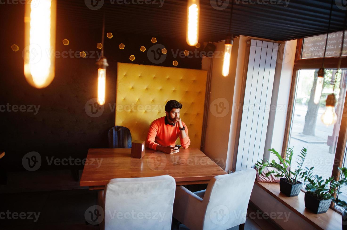 zelfverzekerde jonge indiase man in slimme vrijetijdskleding zoals een oranje trui, zittend in een café en zijn mobiele telefoon gebruikend. foto