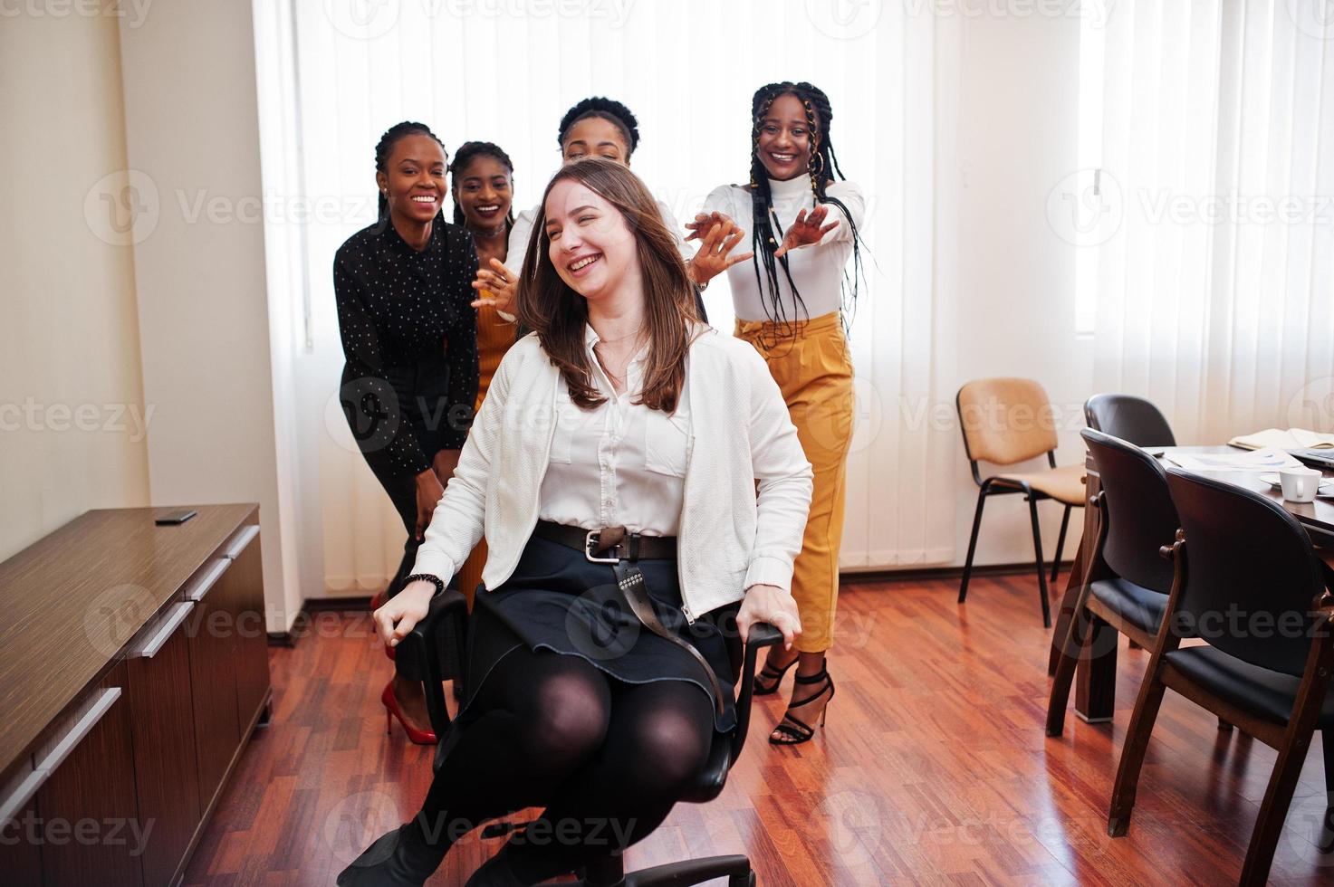 vijf multiraciale zakelijke dames staan op kantoor en rollen vrouw op stoel. diverse groep vrouwelijke werknemers in formele kleding die plezier hebben. foto