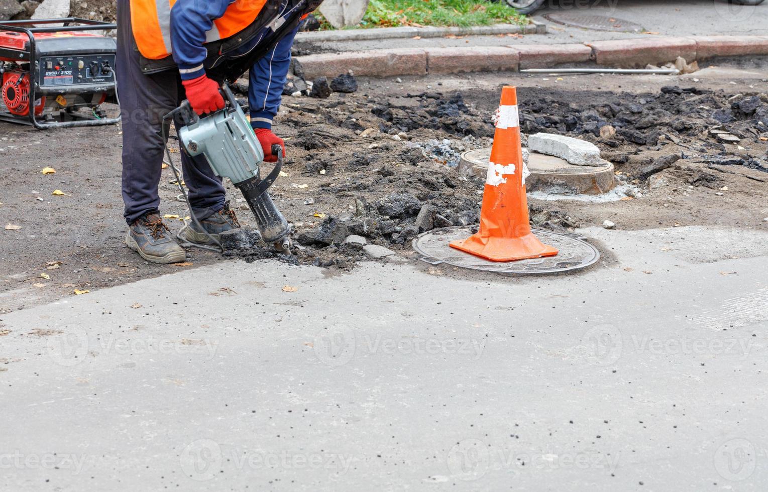 een arbeider met een elektrische drilboor breekt het oude asfalt van de weg rond het rioolmangat. foto