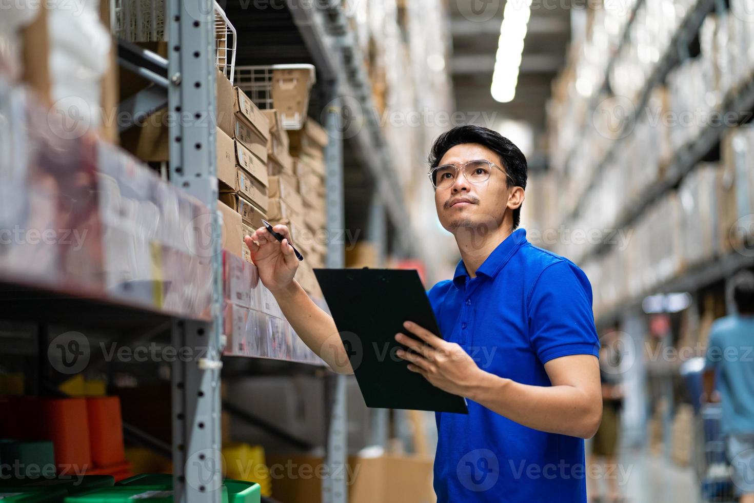 Aziatische mannelijke werknemer in veiligheidspak met tabletcomputer op zoek naar items in een groot magazijn. foto