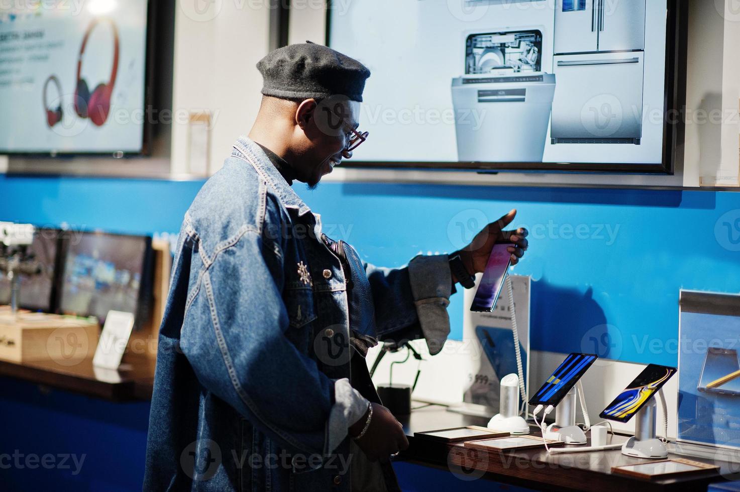 stijlvolle casual Afro-Amerikaanse man bij jeans jasje en zwarte baret met behulp van mobiele telefoon bij elektronicawinkel. foto