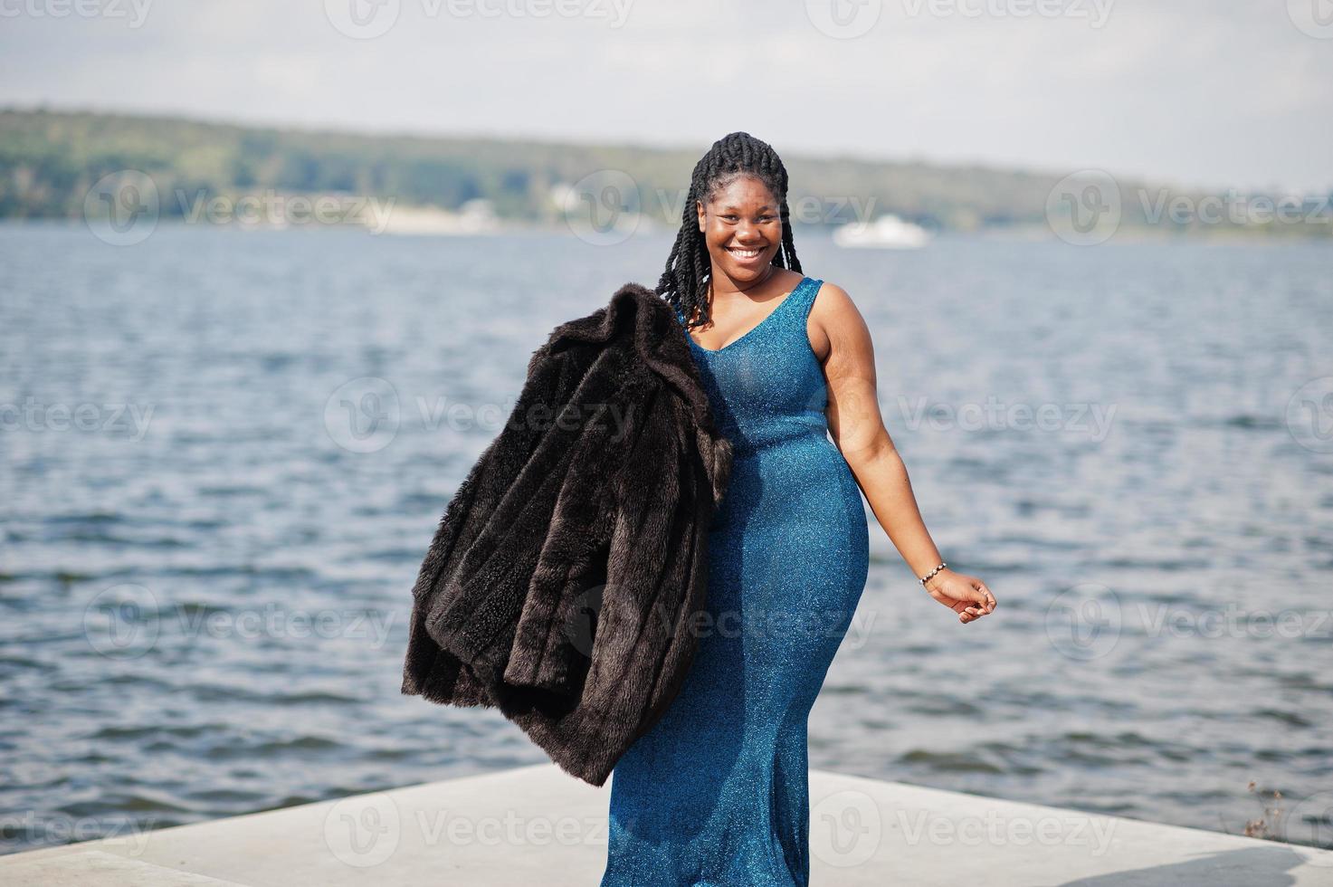 Afro-Amerikaanse donkere huid plus size model poseerde in een blauwe glanzende jurk en zwarte bontjas tegen de zeezijde. foto