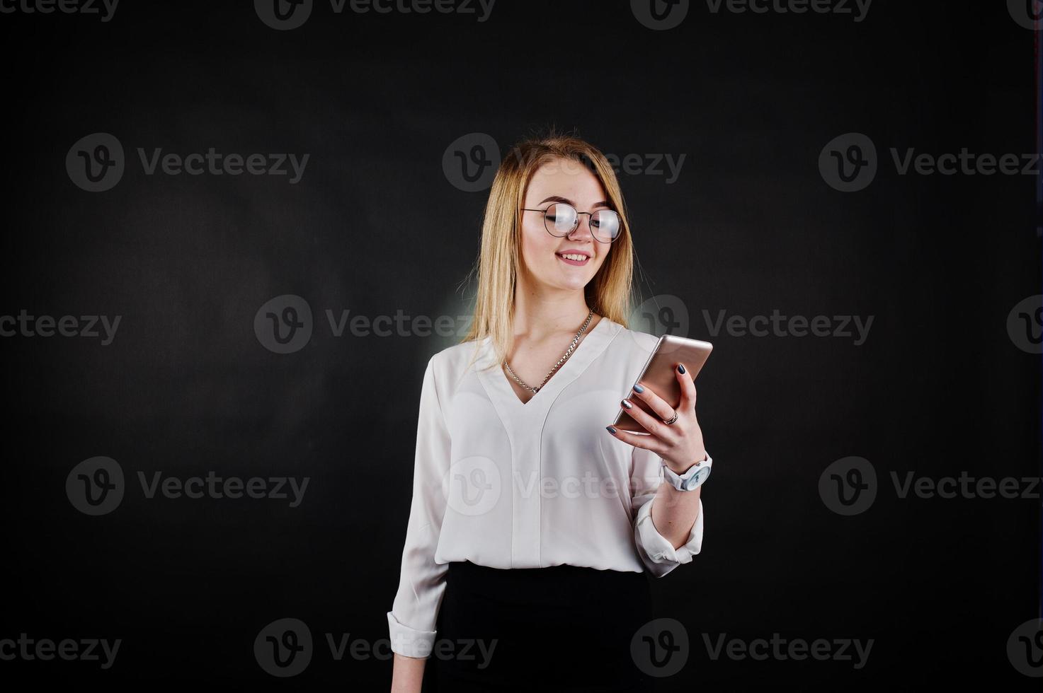 studio portret van blonde zakenvrouw in glazen, witte blouse en zwarte rok kijken naar mobiele telefoon tegen een donkere achtergrond. succesvolle vrouw en stijlvol meisje concept. foto