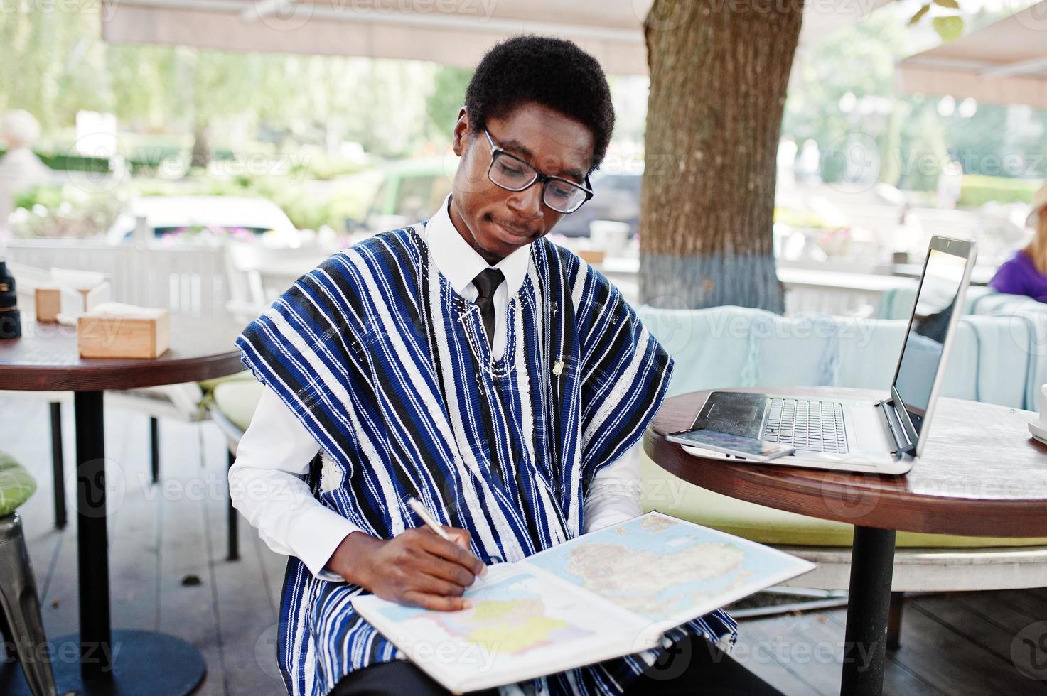 afrikaanse man in traditionele kleding en bril die achter een laptop zit in een buitencafé en op de kaart van afrika en ghana naar zijn notitieboekje kijkt. foto