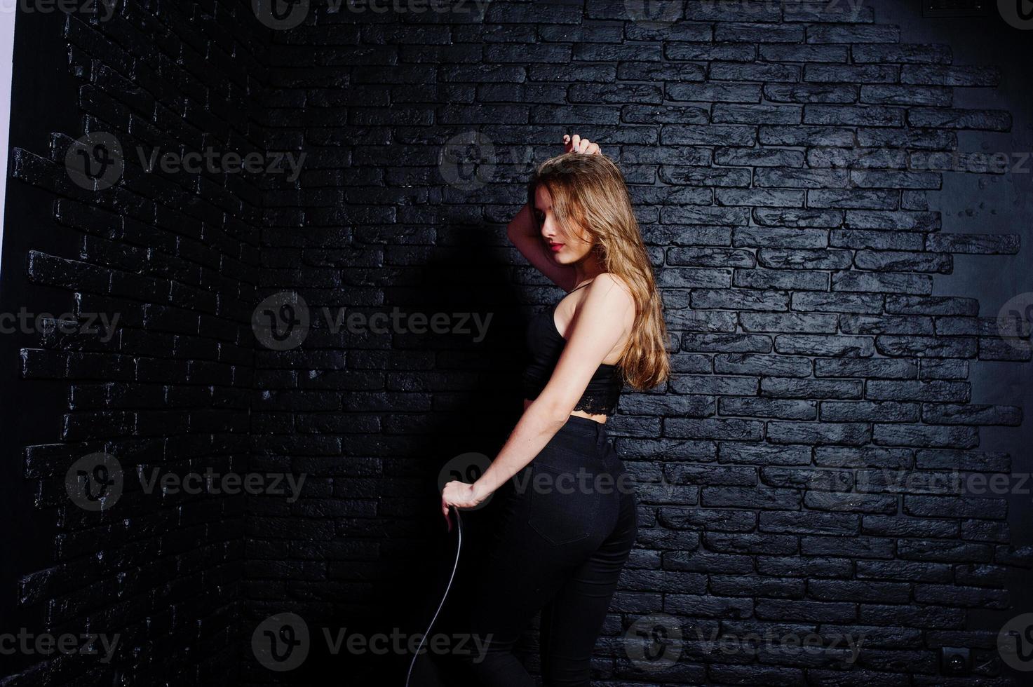 knappe brunette meisje slijtage op zwart, zittend en poseren op stoel in studio tegen donkere bakstenen muur. studiomodel portret. foto