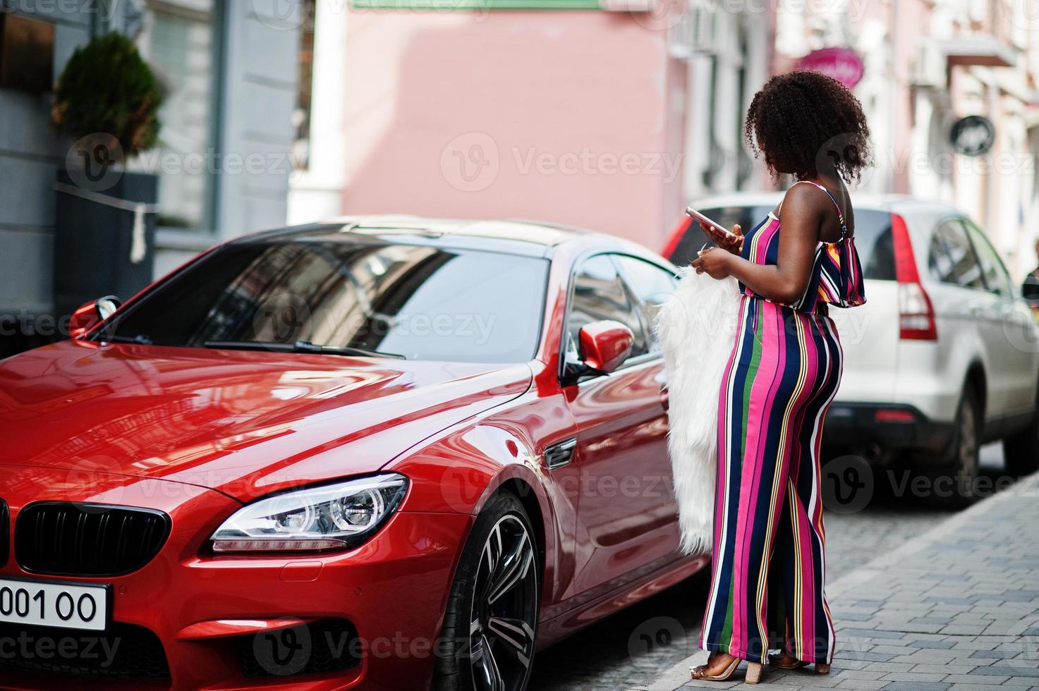 modieuze afro-amerikaanse vrouw in roze gestreepte jumpsuit met pluizige namaakbontjas poseerde tegen rijke rode luxeauto met mobiele telefoon bij de hand. foto